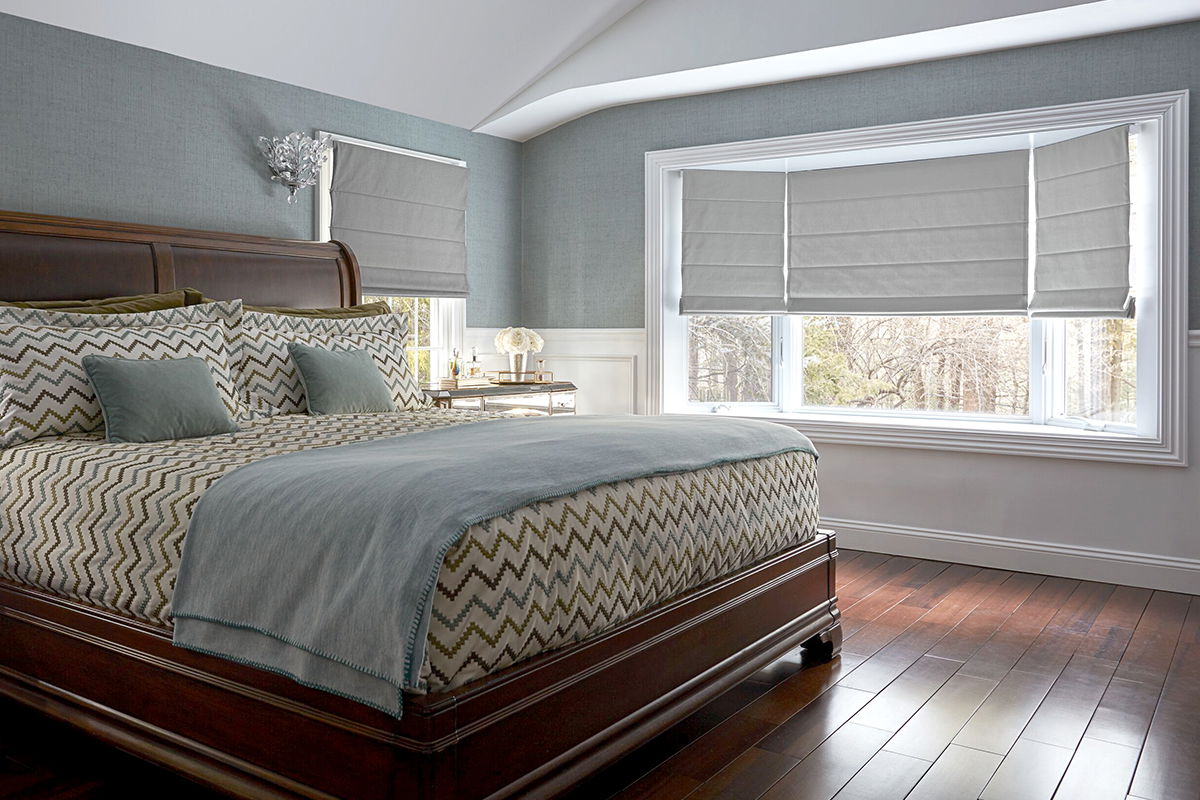 Grey flat Roman shades cover multiple windows, including a bay window, in a spacious bedroom.