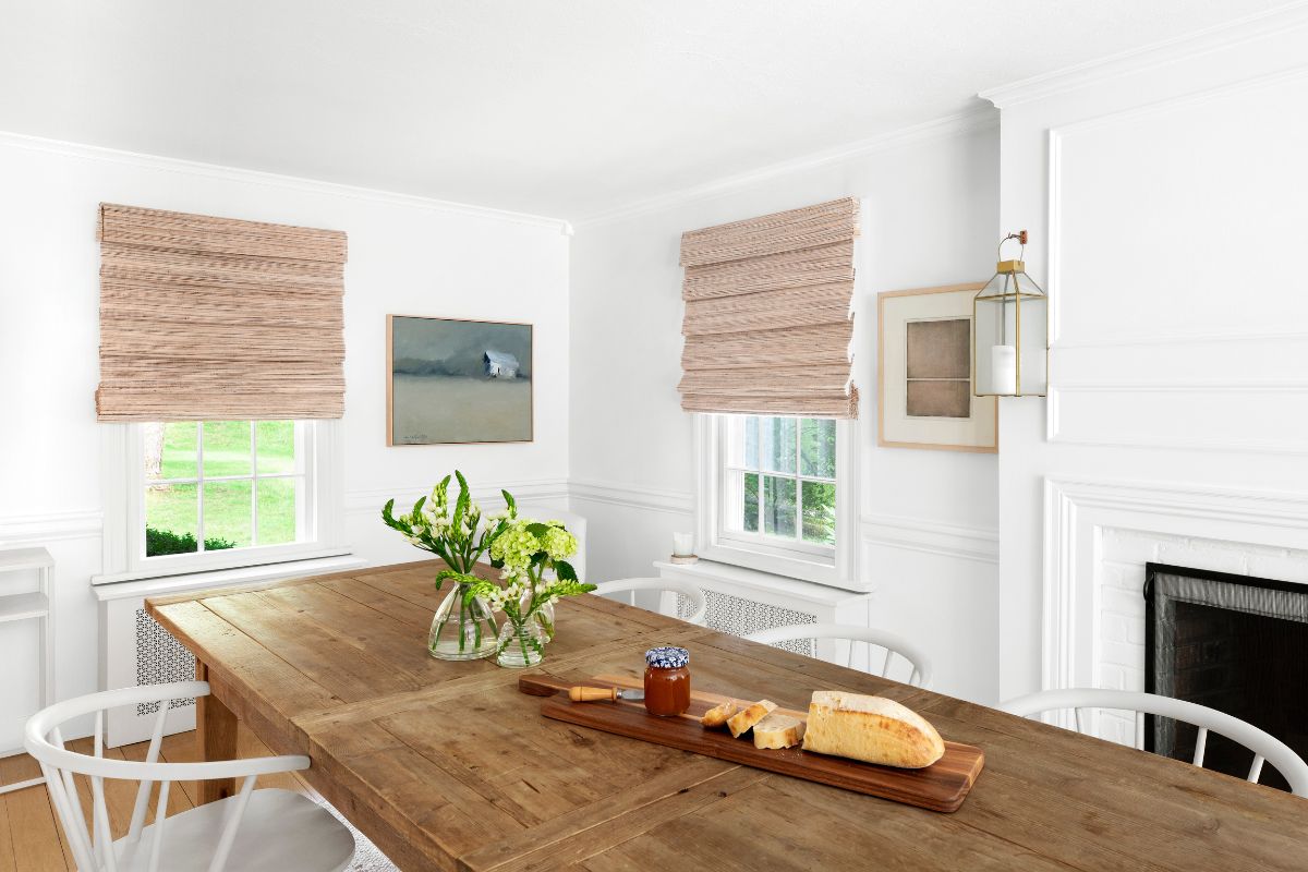 A large rustic dining table is positioned in from of woven wood shades on two windows.