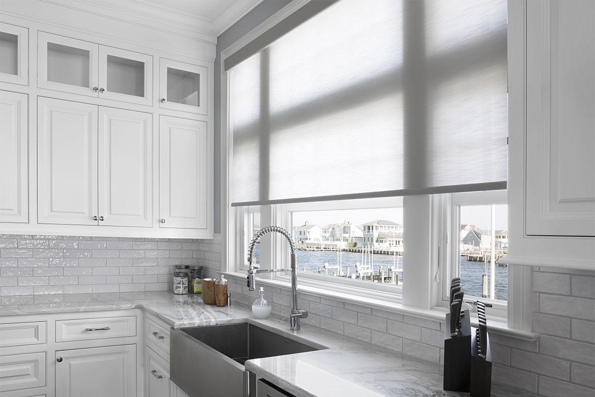 A white roller shade covers a large kitchen window with a costal view.
