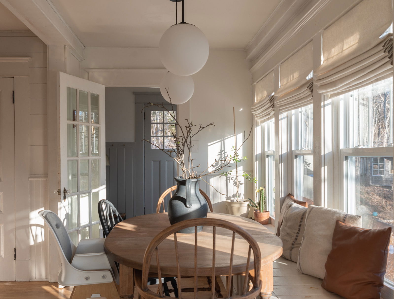 Natural color Roman shades cover three windows in a sun room with a farmhouse dining table.