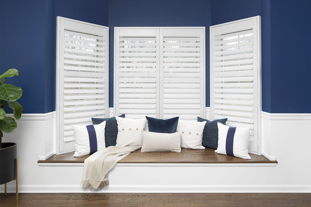 White indoor shutters are slightly ajar covering a large bay window with a sitting area.