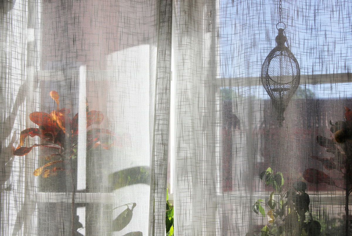 Sheer white linen drapes allow the sun to pour in from a window with many plants on the window sill.